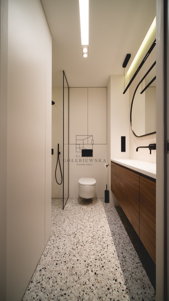 Nowoczesna łazienka z kabiną prysznicową typu walk-in, umywalką z drewnianą szafką, okrągłym lustrem z podświetleniem LED i podłogą z lastryko w odcieniach szarości i bieli.