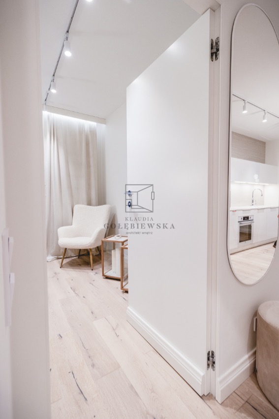 Jasne wnętrze z widokiem na salonik z kremowym fotelem i stolikiem, oraz kuchnię odbijającą się w owalnym lustrze, całość utrzymana w bieli i jasnych odcieniach drewna.