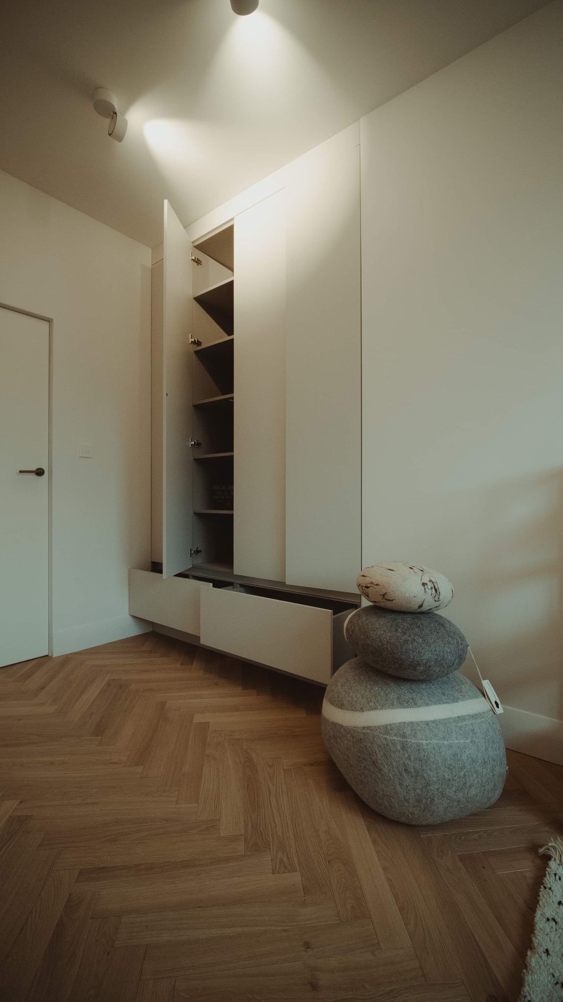 Minimalistyczne wnętrze z zabudową meblową w odcieniach bieli, drewnianą podłogą w jodełkę i dekoracyjnymi poduszkami ułożonymi w stos. Otwarta szafka z półkami ukazuje przechowywane przedmioty.