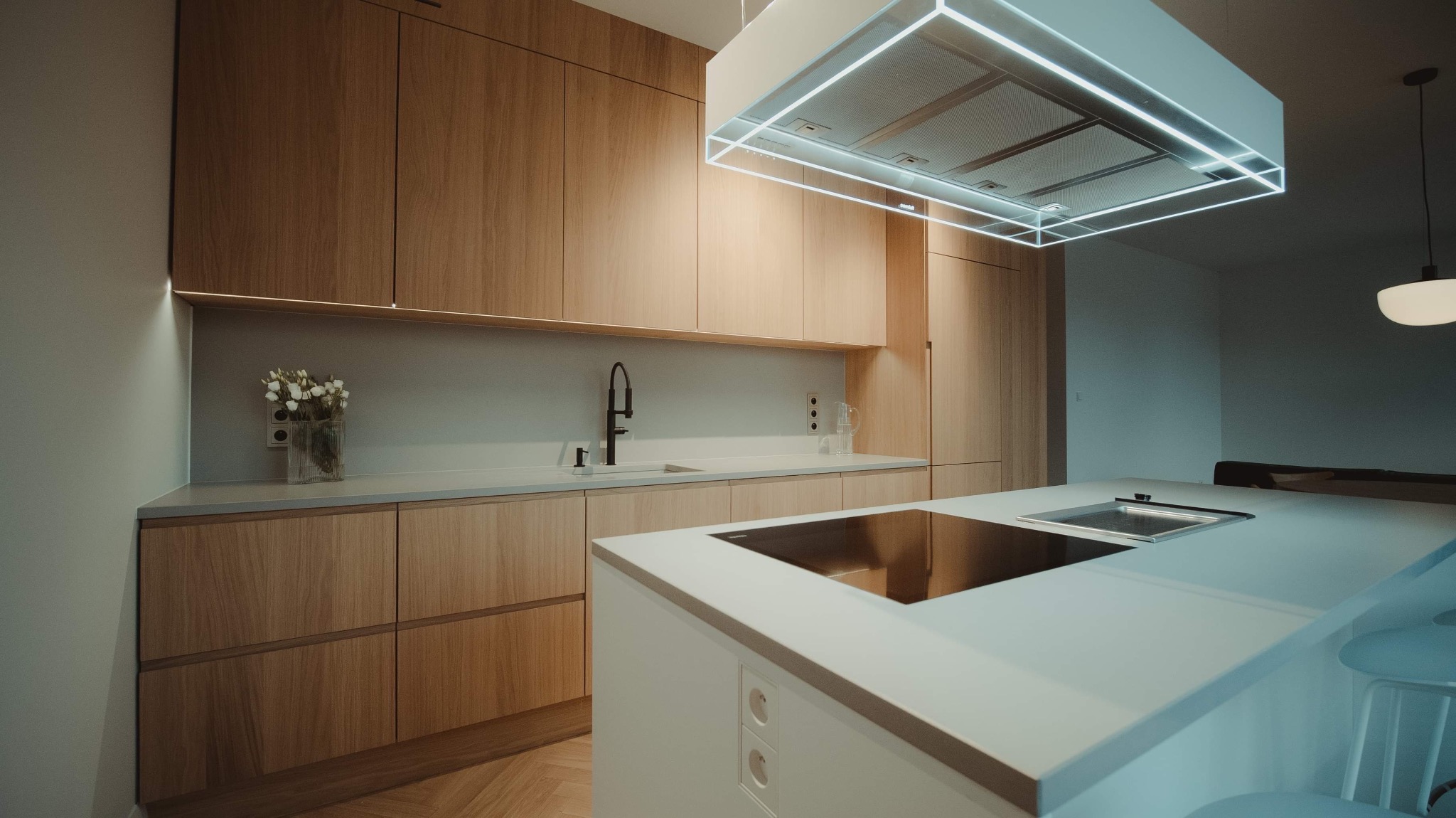 Nowoczesna kuchnia z drewnianymi frontami szafek, białym blatem i wyspą, oświetlona lampą sufitową z neonowym obramowaniem i wiszącą lampą.