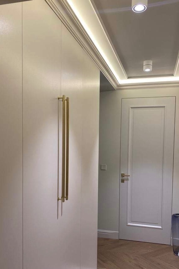 Minimalistyczna szafa wnękowa z długimi, złotymi uchwytami na tle białych drzwi i parkietu ułożonego w jodełkę, oświetlona punktowymi lampami sufitowymi i listwą LED.