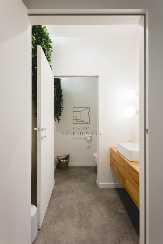 Nowoczesna łazienka z betonową podłogą, wiszącą zielenią i drewnianą szafką pod umywalką, widziana zza uchylonych drzwi. Logo projektanta wnętrz na ścianie.