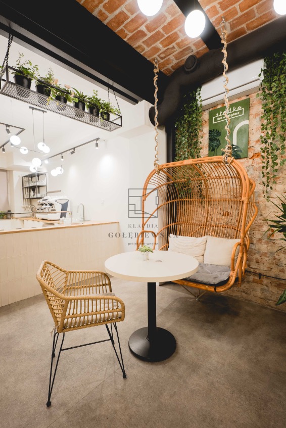 Wnętrze kawiarni z ceglaną ścianą, wiszącym fotelem z wikliny na linach, okrągłym stolikiem z czarną nogą i wiklinowym krzesłem na metalowych nogach, podwieszane rośliny w czarnych doniczkach.