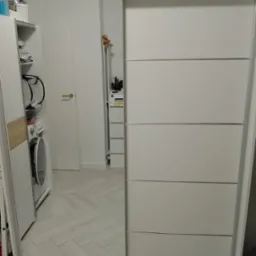 Biała szafa przesuwna z lustrem, w którym odbija się wnętrze pokoju z pralką i białymi meblami.