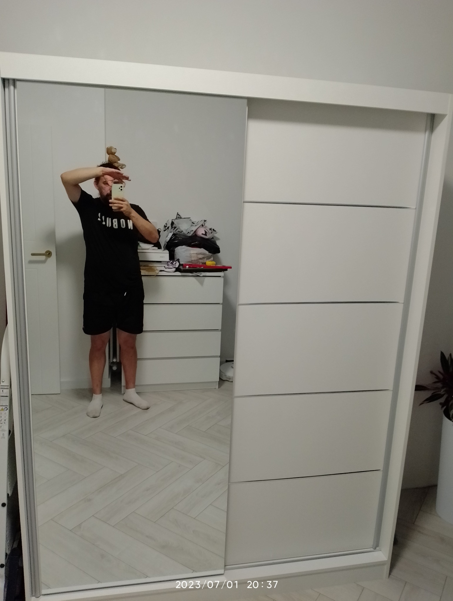 Mężczyzna w czarnym stroju robi selfie w lustrze szafy przesuwnej z białymi frontami na tle komody i podłogi w jodełkę, z pluszowym misiem na głowie.