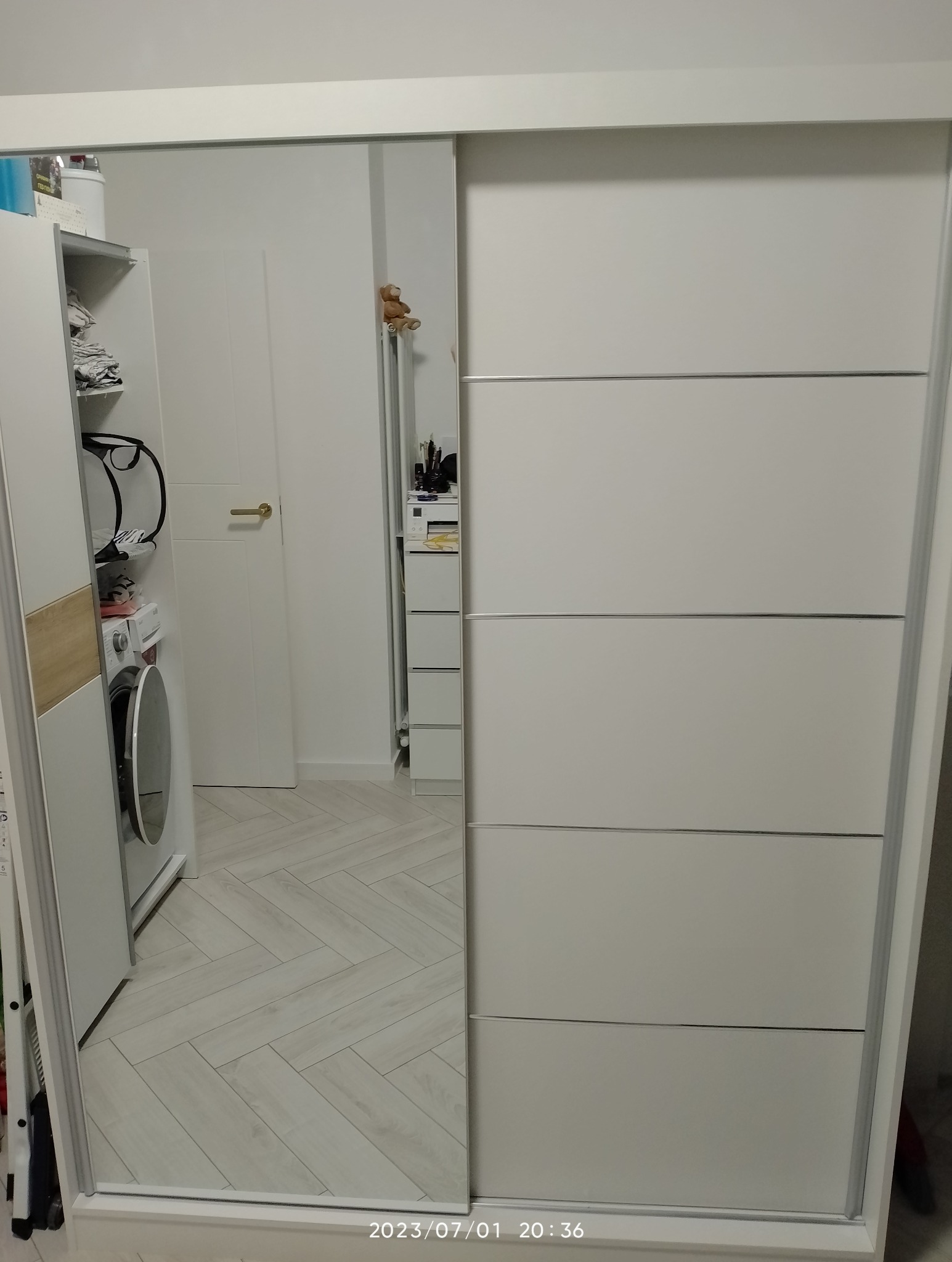 Biała szafa przesuwna z lustrem, w którym odbija się wnętrze pokoju z pralką i białymi meblami.