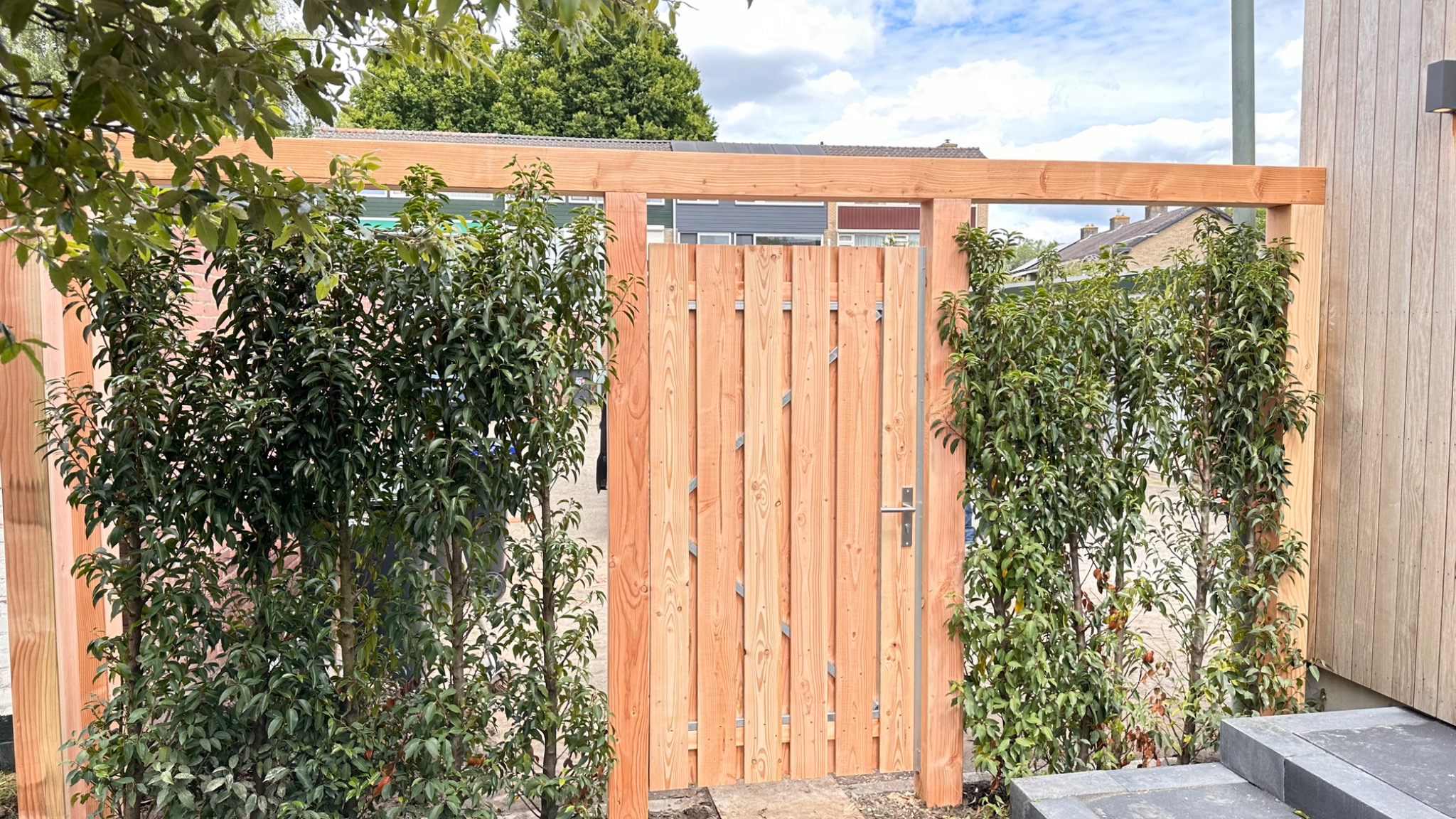 Nowoczesna drewniana furtka z pionowymi deskami w ogrodzeniu, otoczona zielenią, z widocznymi elementami konstrukcyjnymi i metalową klamką.