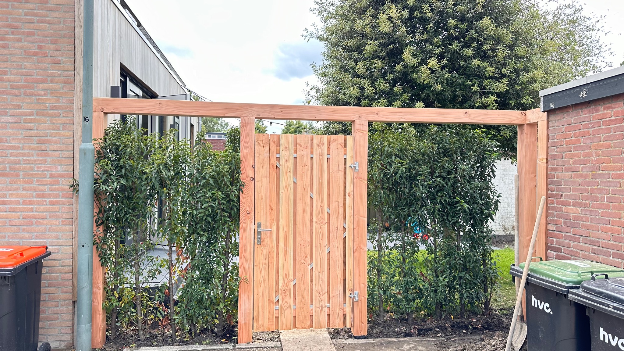 Nowa drewniana furtka z ramą, wkomponowana w żywopłot, widok z zewnątrz, z widocznymi koszami na śmieci po bokach.