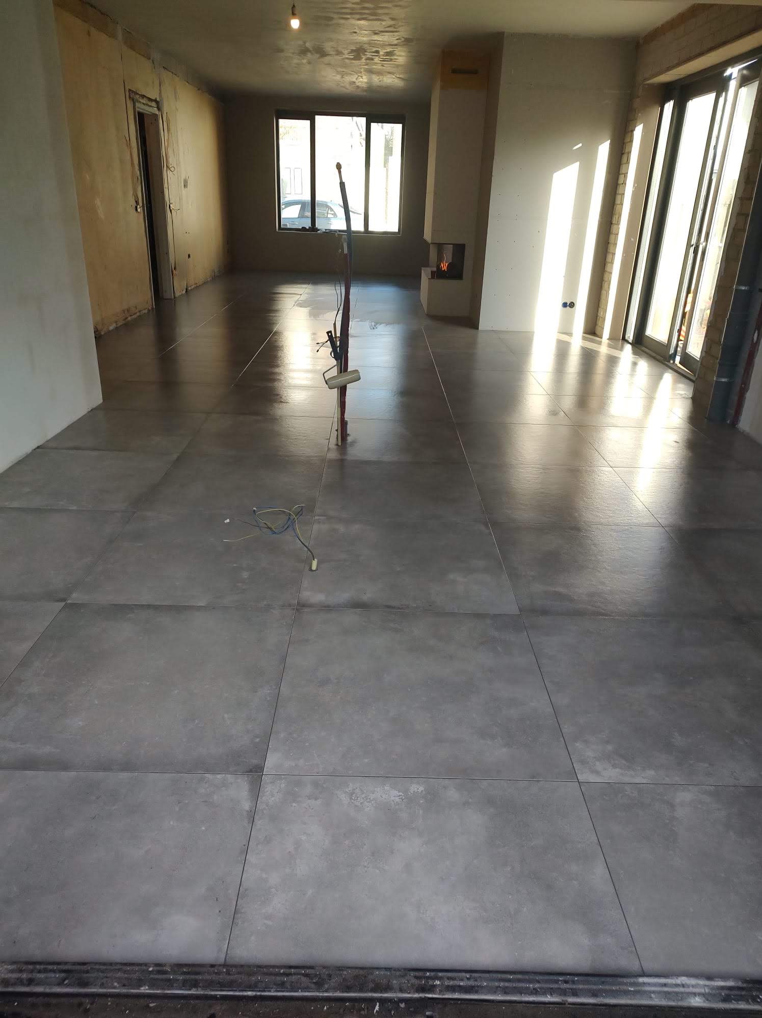 Wnętrze w trakcie wykańczania z dużymi szarymi płytkami podłogowymi, widocznymi przewodami, kominkiem i dużymi oknami wpuszczającymi jasne światło.