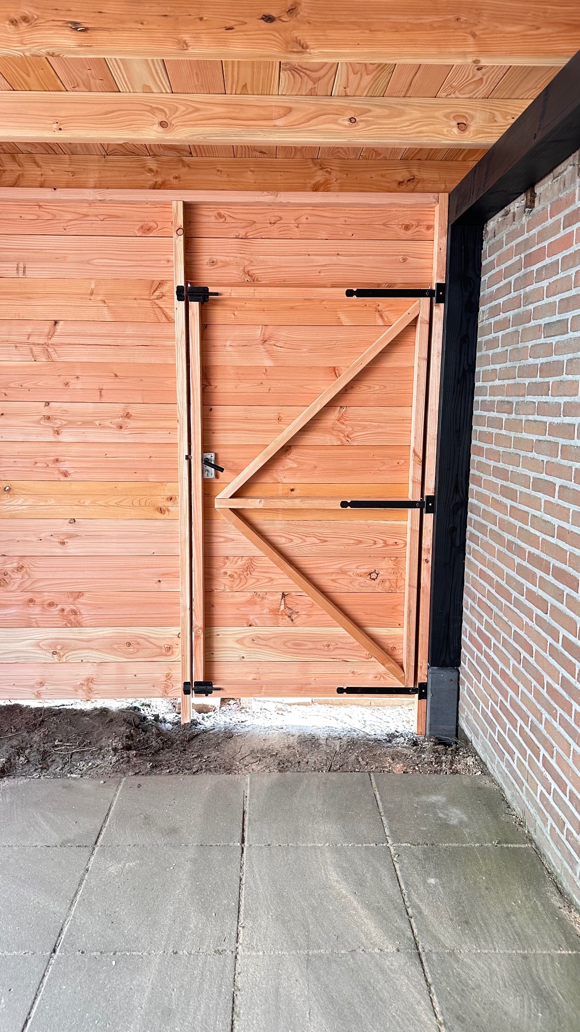 Wykończona drewniana brama z czarnymi zawiasami, wbudowana w drewnianą konstrukcję z ceglanym murem z boku, widoczna kostka brukowa i fragment ziemi.