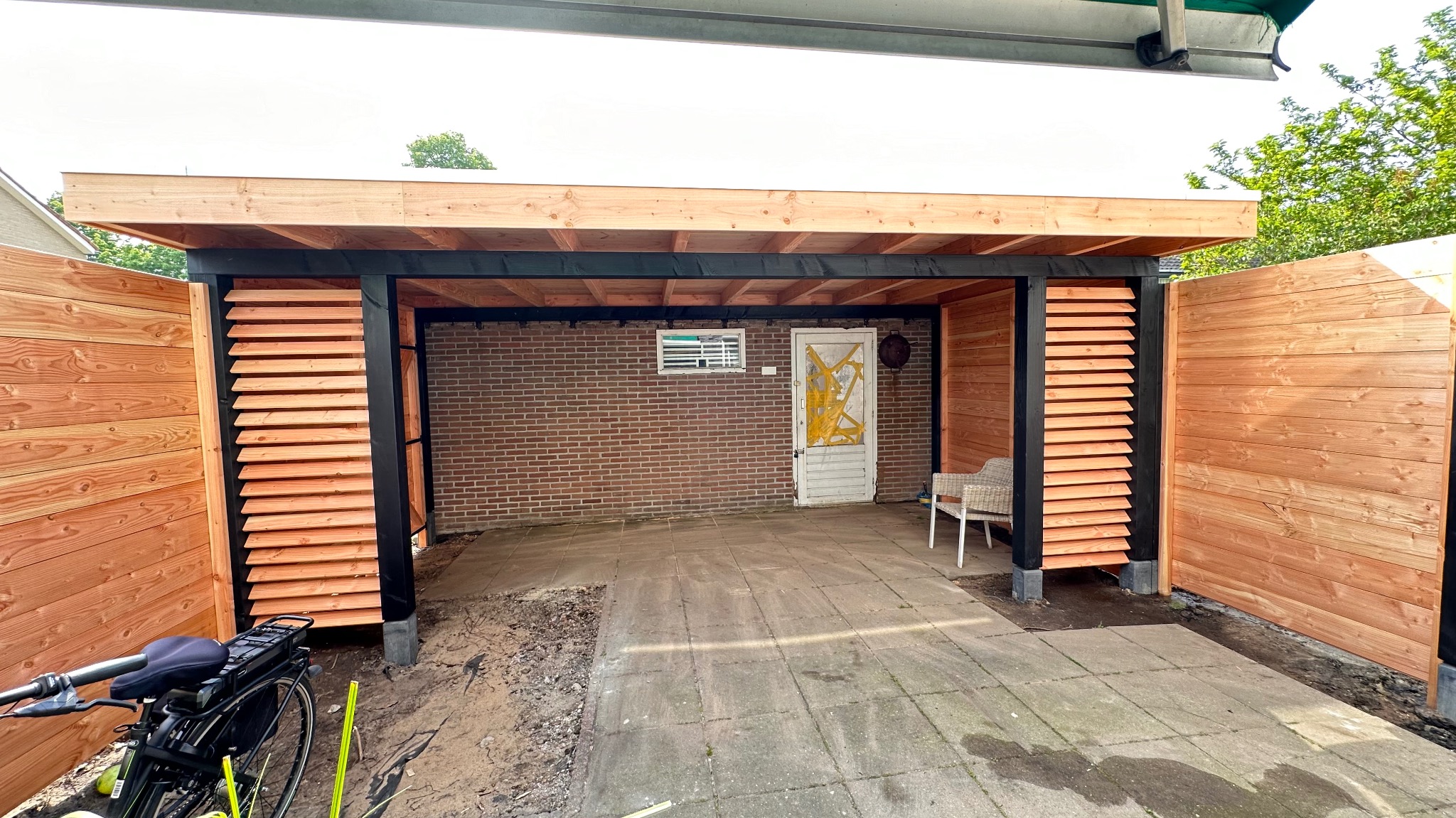 Nowoczesna wiata garażowa z drewnianą zabudową i żaluzjami, rower elektryczny zaparkowany z boku, widoczna ceglana ściana z oknem i starą, oklejoną taśmą, białą furtką.