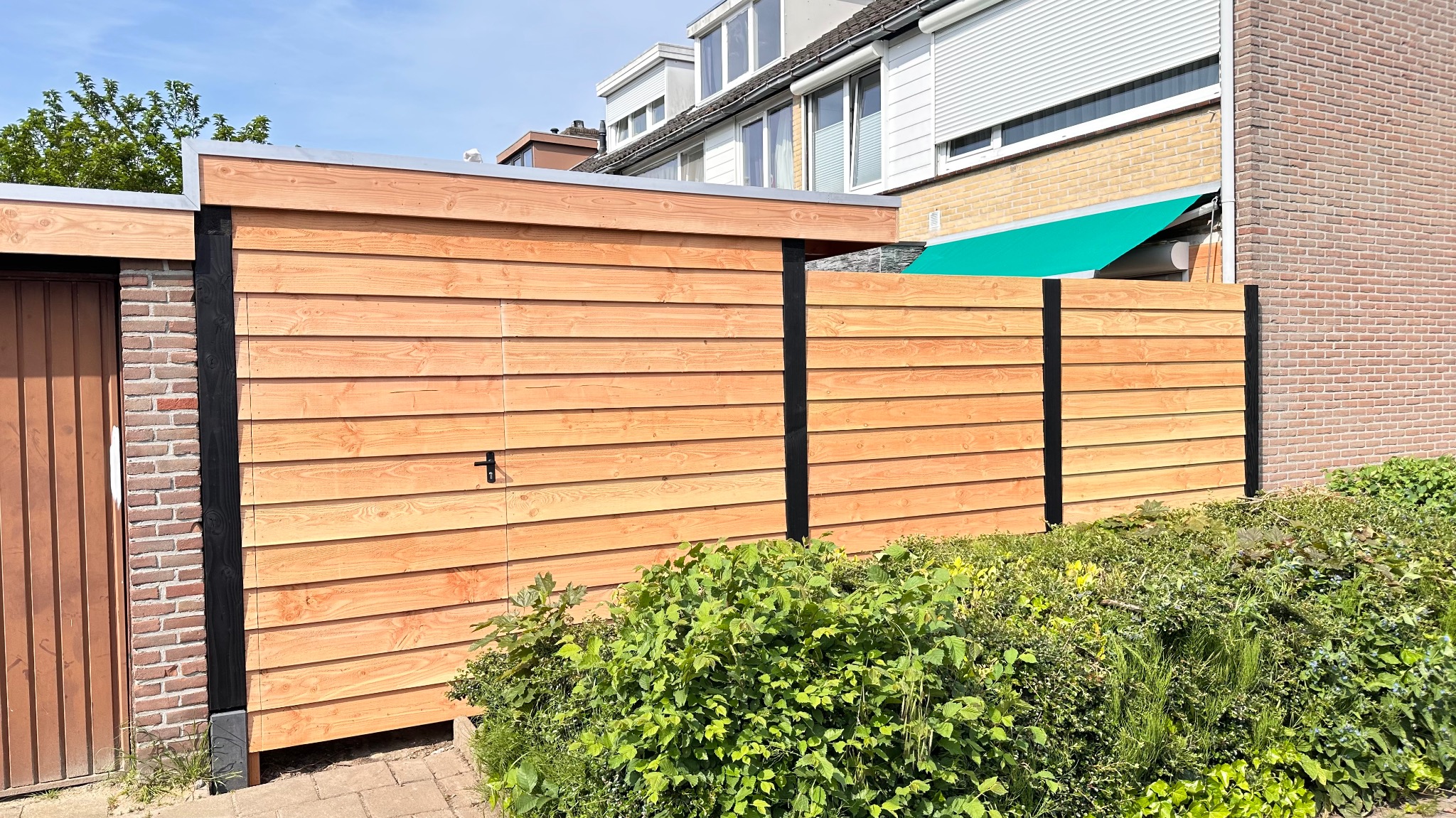 Nowoczesna drewniana zabudowa ogrodowa z poziomych desek, z czarnymi słupkami, przylegająca do ceglanego budynku i bujnej zieleni.