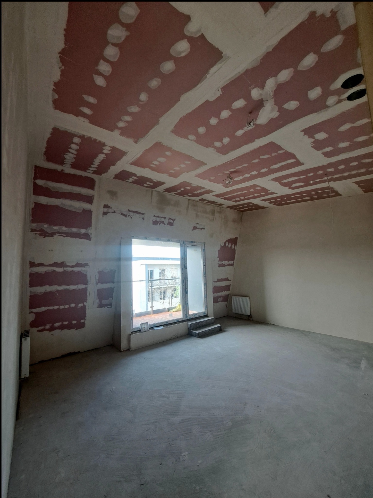 Wnętrze w trakcie prac wykończeniowych: surowy beton na podłodze, ściany z fragmentami płyt gips-karton i szpachli, przygotowane do dalszych prac. Widoczne okno i grzejnik.