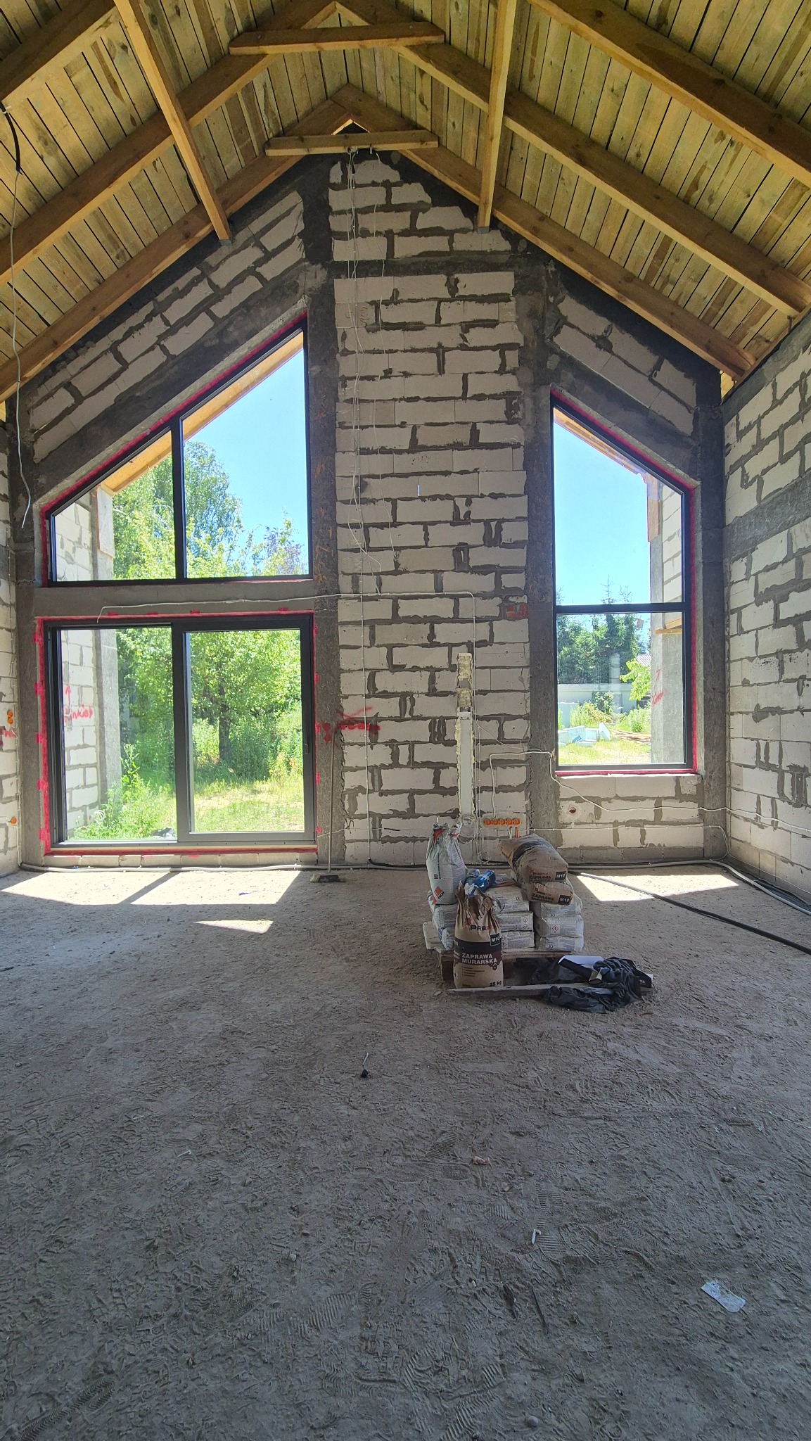 Wnętrze domu w trakcie budowy: surowe ściany z betonu komórkowego, duże okna z widokiem na zieleń, drewniana konstrukcja dachu. Widoczne worki z zaprawą murarską.