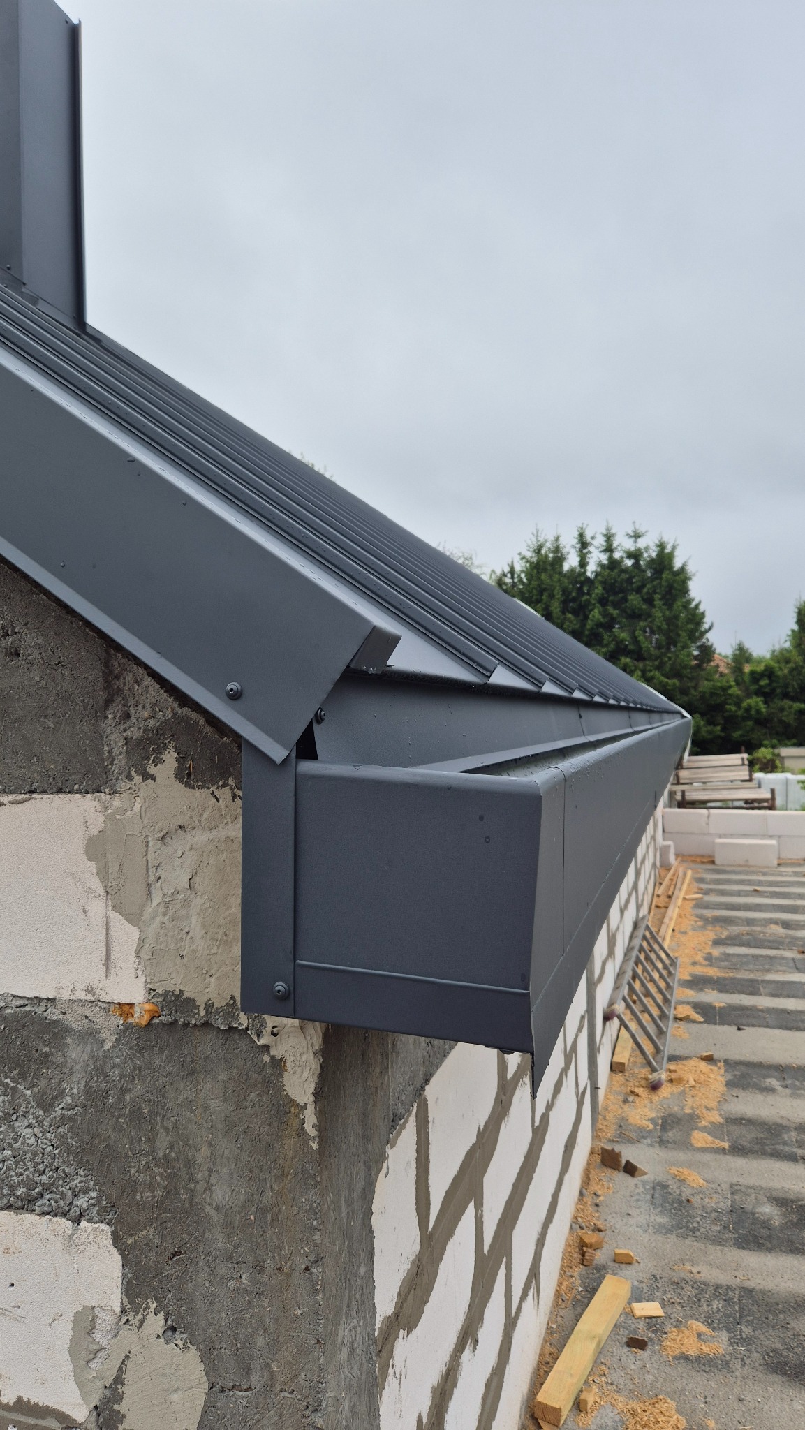 Szary, metalowy system orynnowania na murowanej ścianie domu w trakcie budowy, widok z dołu, na tle zieleni.