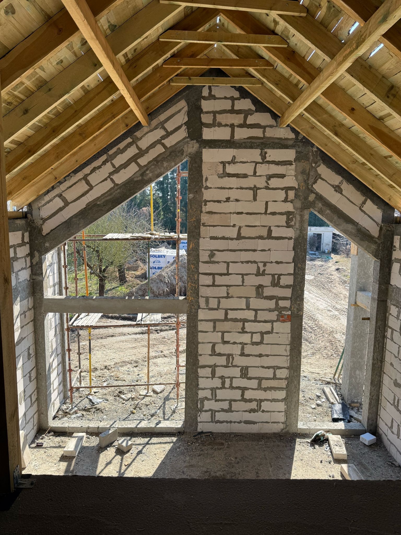 Wnętrze domu w trakcie budowy: surowe ściany z betonu komórkowego, konstrukcja dachu z drewna, widok na zewnątrz przez otwory okienne. W tle rusztowanie.