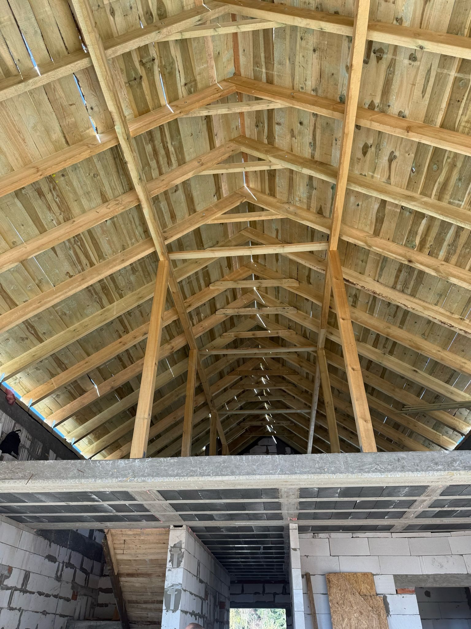 Wnętrze w trakcie budowy: drewniana konstrukcja dachu na tle surowych ścian z betonu komórkowego i stropu. Widok z dołu ukazuje skomplikowaną sieć belek.