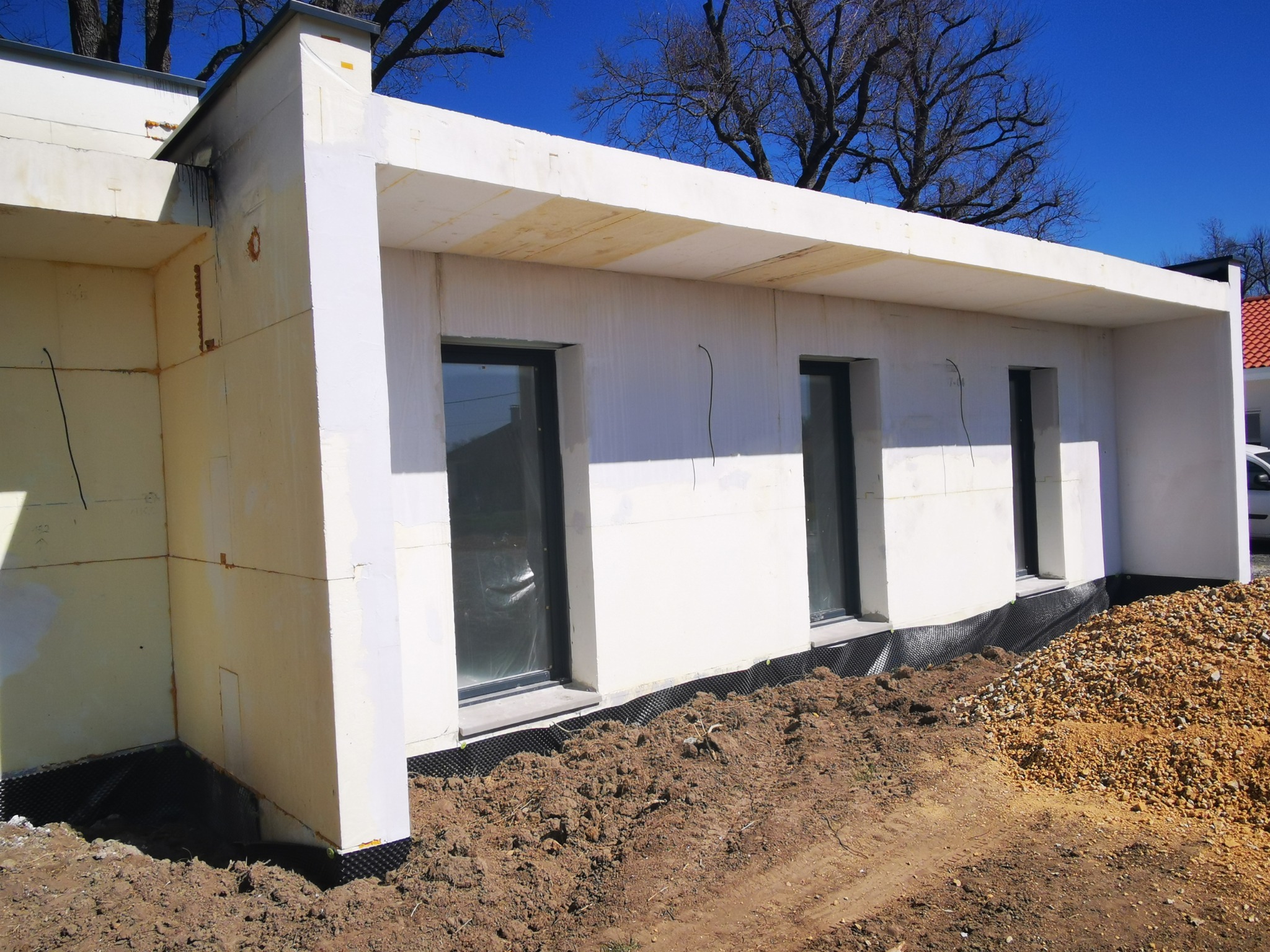 Surowy stan domu modułowego z białymi ścianami, widoczną izolacją i czarną folią fundamentową. Okna zabezpieczone folią, ziemia i kamienie wokół.