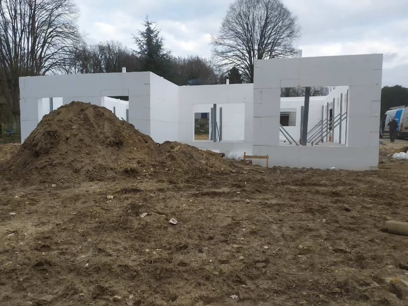 Budowa domu z białych paneli, widoczny stos ziemi i metalowe wsporniki. Ujęcie z zewnątrz, w tle drzewa i częściowo widoczny samochód dostawczy.