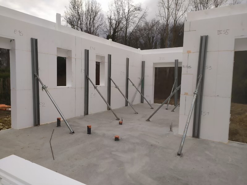 Wnętrze domu w trakcie budowy z białych bloków styropianowych, wzmocnionych metalowymi szynami i podporami. Widoczne otwory okienne i drzwiowe, surowy beton na podłodze.