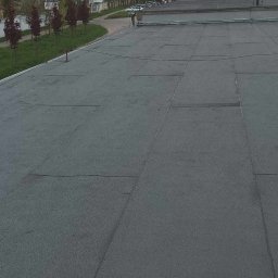 Aesthetics - Szary, płaski dach pokryty papą termozgrzewalną z widocznymi łączeniami, w tle budynki mieszkalne i przemysłowe, drzewa i słupy oświetleniowe.