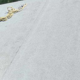 Aesthetics - Szary, płaski dach pokryty papą z widocznymi łączeniami, z żółtą liną zabezpieczającą i fragmentem drzewa w tle.