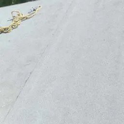 Szary, płaski dach pokryty papą z widocznymi łączeniami, z żółtą liną zabezpieczającą i fragmentem drzewa w tle.