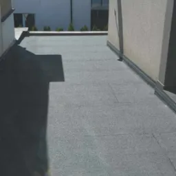 Płaski dach pokryty papą termozgrzewalną w kolorze szarym, widoczne łączenia arkuszy, obróbka blacharska przy krawędzi, w tle budynek mieszkalny z anteną satelitarną.