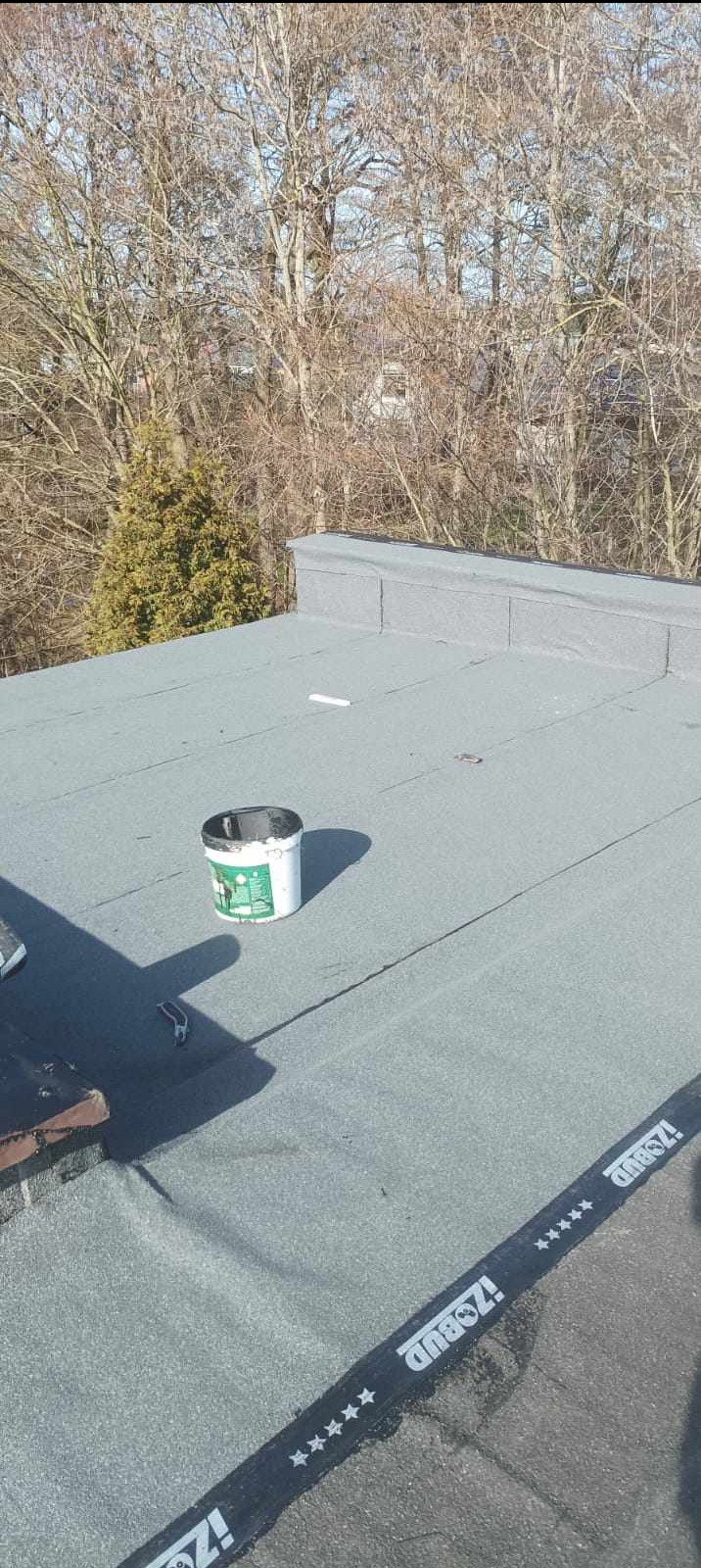 Szary dach pokryty papą z widoczną puszką farby, fragmentem narzędzia i taśmą uszczelniającą z napisem 'IZOBUD' oraz drzewami w tle.