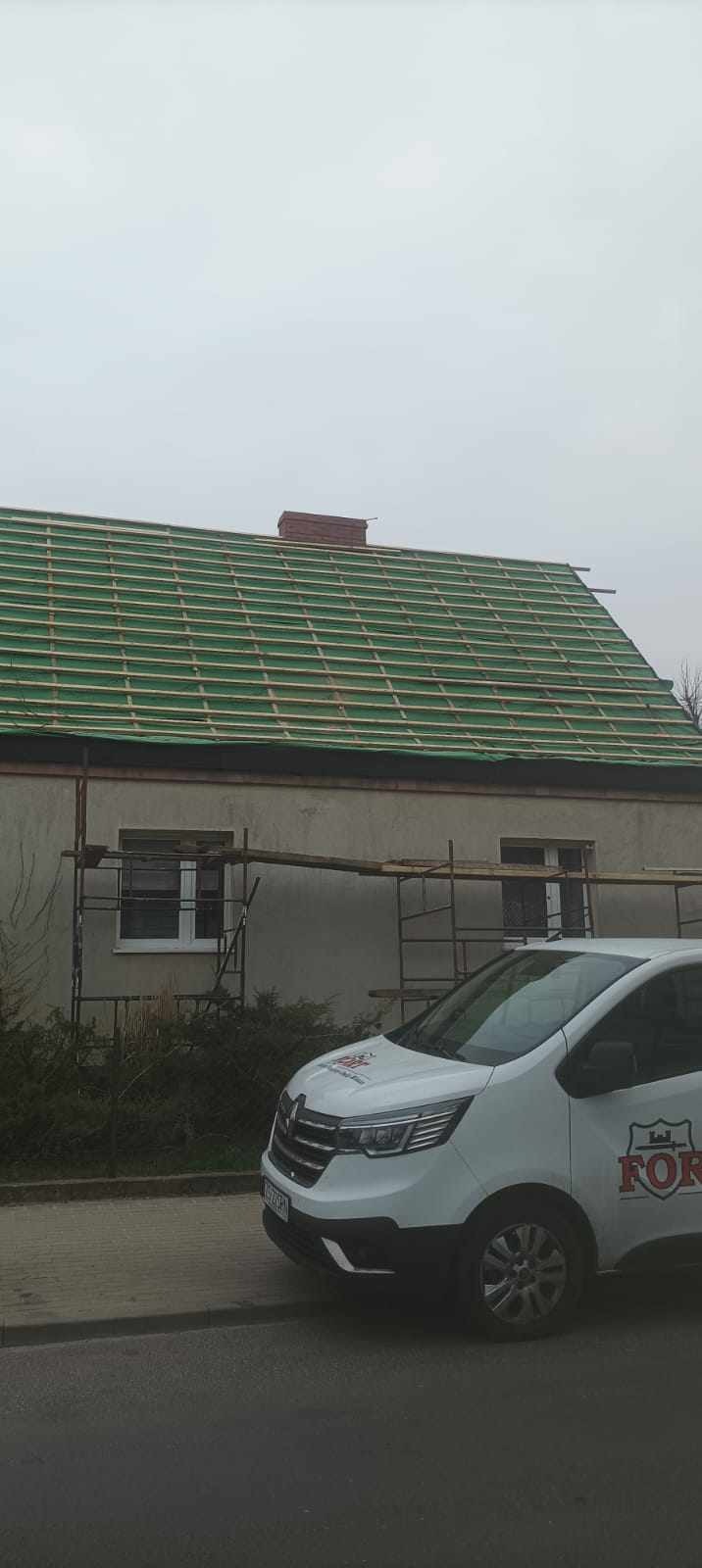 Dom w trakcie przygotowywania do pokrycia dachu, widoczne łaty i kontrłaty, rusztowanie oraz samochód dostawczy z logo firmy.