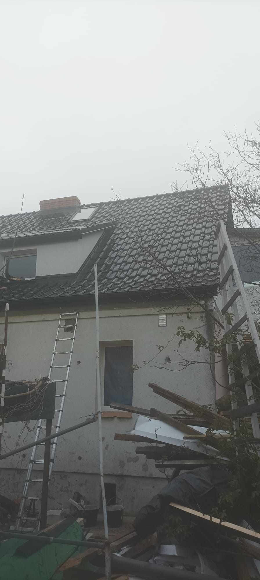 Dom z ciemną dachówką, widoczne dwie drabiny opierające się o ścianę, w tle mgła i drzewa bez liści, na ziemi porozrzucane deski i materiały budowlane.