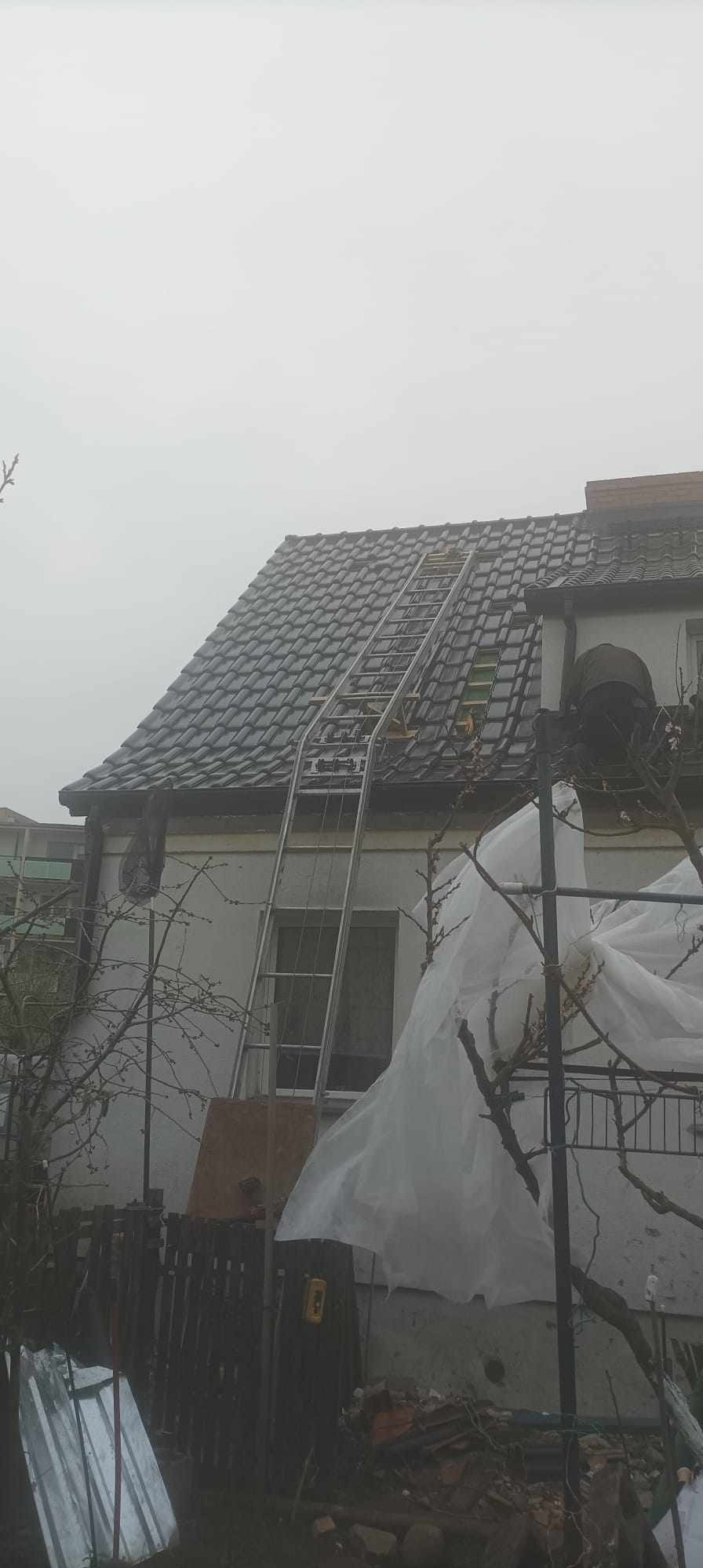 Dom jednorodzinny z ciemną dachówką, na dachu drabina, widoczne prace dekarskie przy krawędzi dachu, pochmurne niebo w tle.
