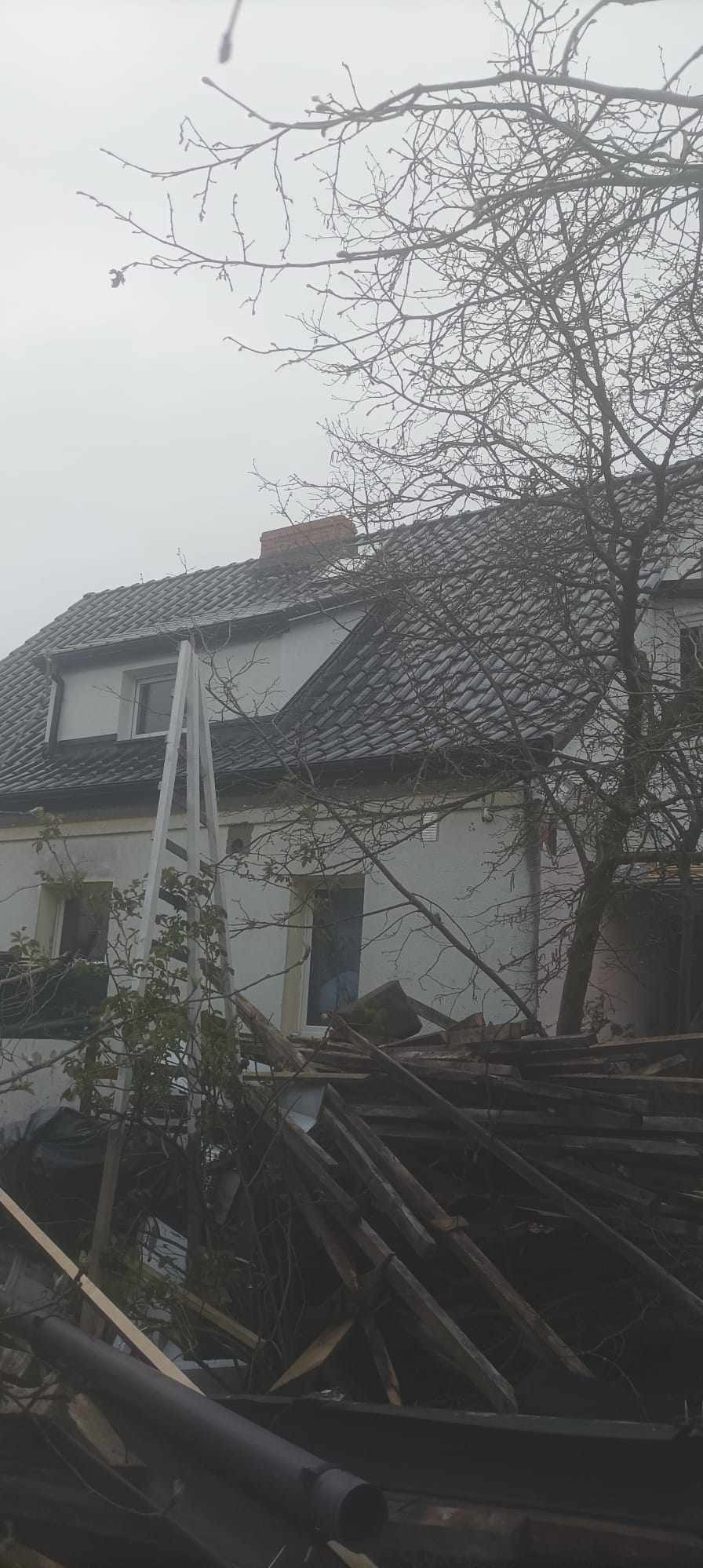 Dom z dachem pokrytym szarą dachówką, widoczne rusztowanie i sterta starych desek na pierwszym planie, pochmurna pogoda.