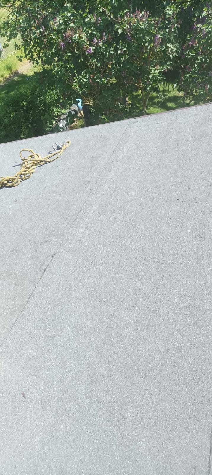 Szary, płaski dach pokryty papą z widocznymi łączeniami, z żółtą liną zabezpieczającą i fragmentem drzewa w tle.