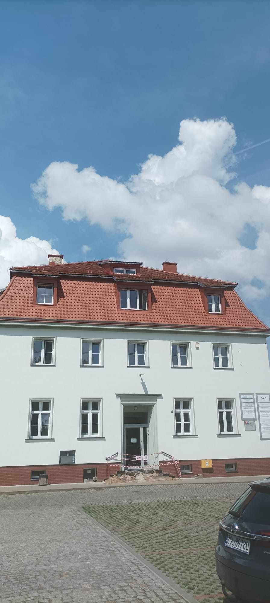 Dwupiętrowy budynek z czerwonym dachem z dachówki ceramicznej, widoczne okna dachowe, elewacja w kolorze złamanej bieli, zabezpieczone wejście taśmą ostrzegawczą.