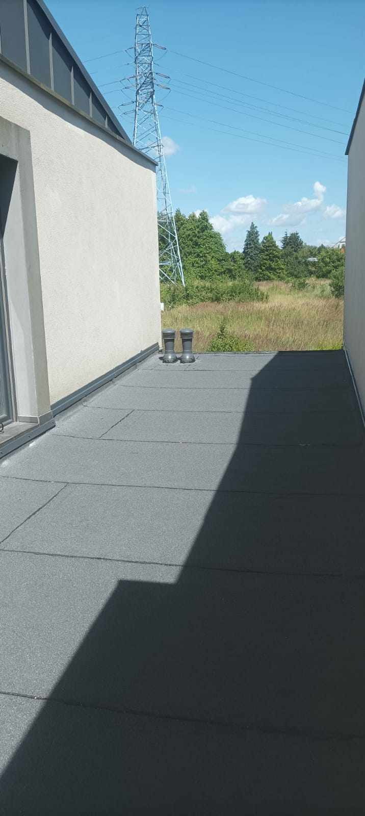 Szary, płaski dach pokryty papą termozgrzewalną, z dwoma kominkami wentylacyjnymi, widok na zewnątrz budynku z słupem wysokiego napięcia w tle.