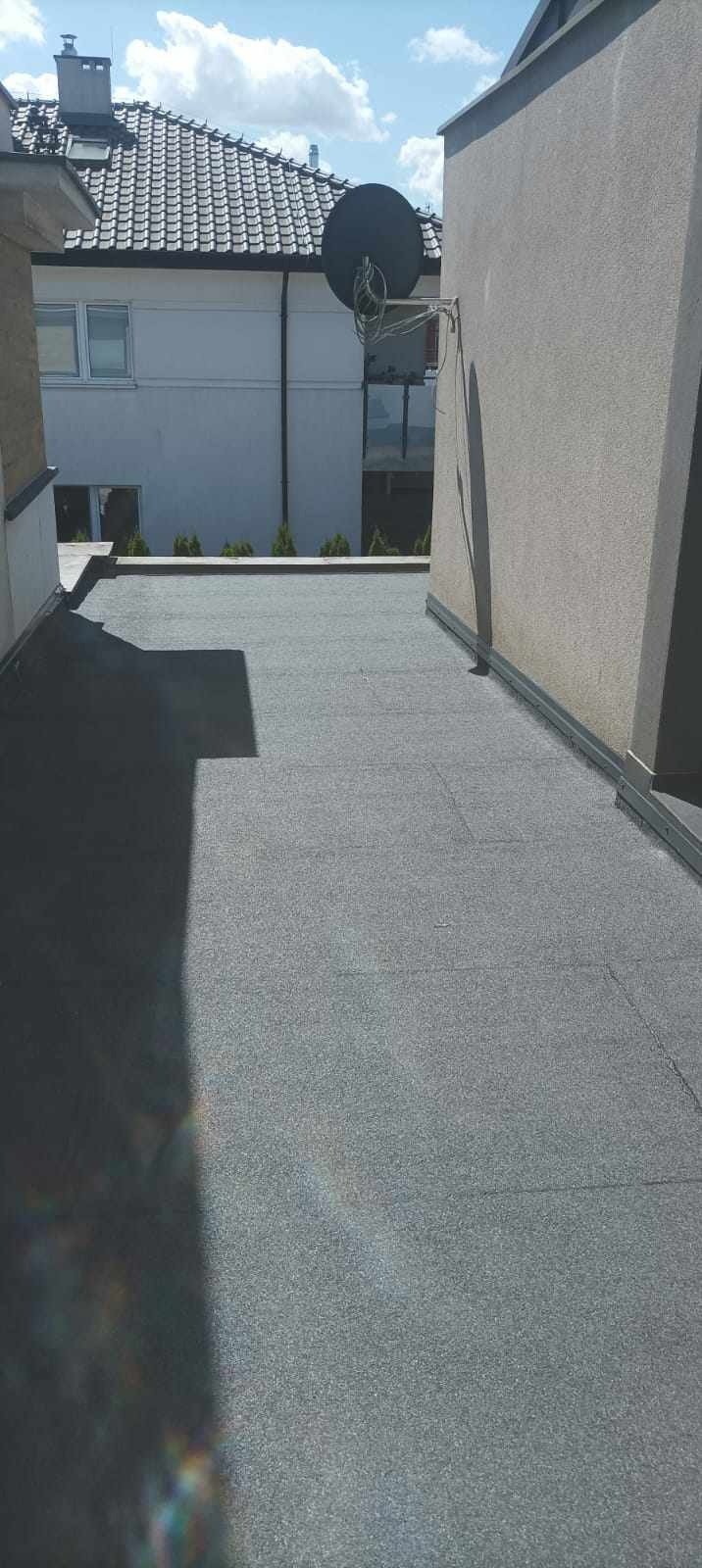 Płaski dach pokryty papą termozgrzewalną w kolorze szarym, widoczne łączenia arkuszy, obróbka blacharska przy krawędzi, w tle budynek mieszkalny z anteną satelitarną.