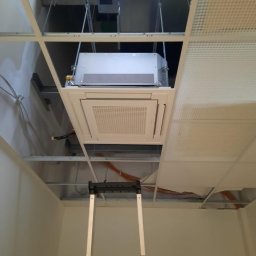 Tomasz Kozik - Montaż klimatyzacji kasetonowej w biurze, widok od dołu z drabiny, instalacja w suficie podwieszanym, widoczne kable i elementy konstrukcyjne sufitu.