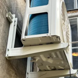 Zewnętrzna jednostka klimatyzatora zamontowana na ścianie budynku, widoczne elementy montażowe i fragment okna w tle.