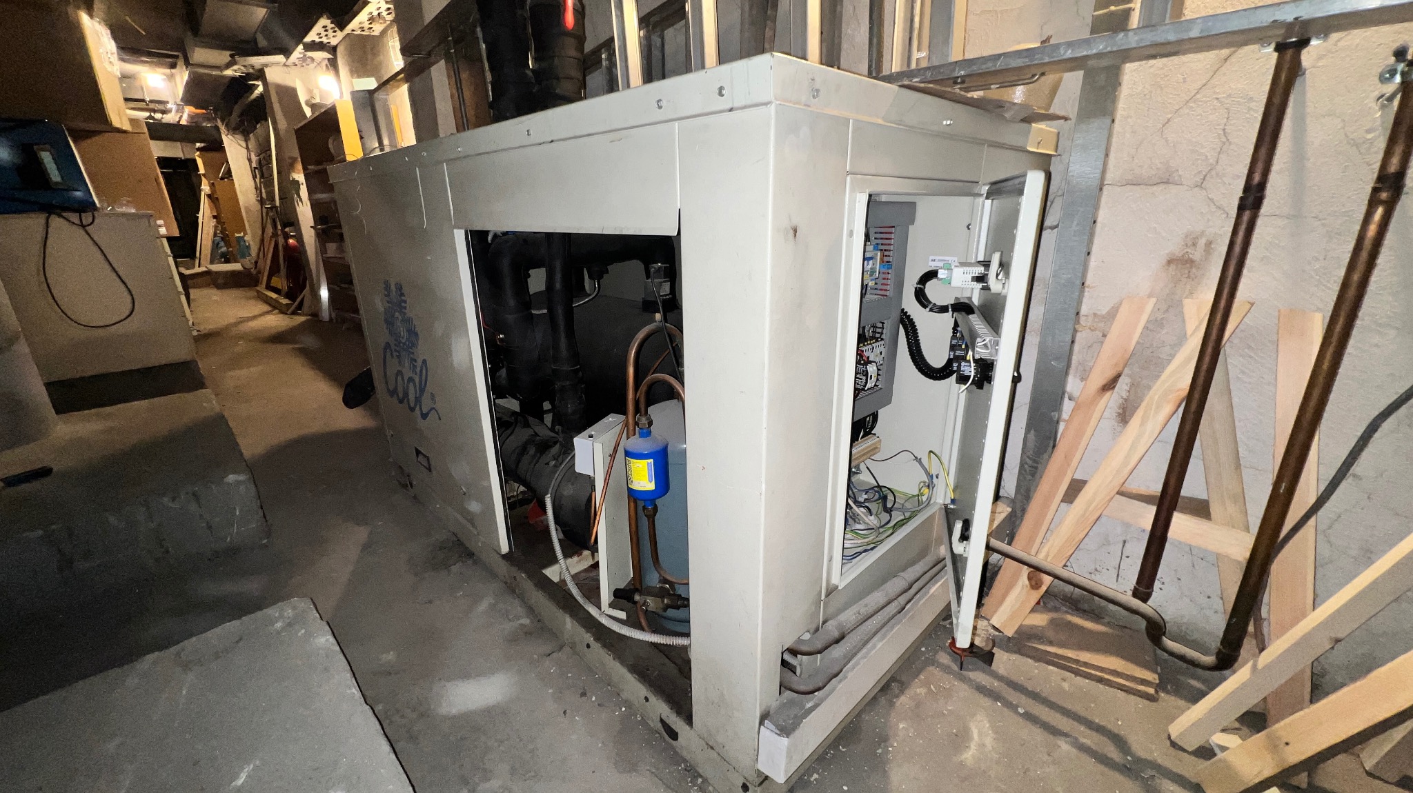 Duża centrala klimatyzacyjna w pomieszczeniu technicznym, widoczne rury miedziane, okablowanie i elementy sterujące w otwartej obudowie, obok drabina i prowizoryczne podpory z drewna.