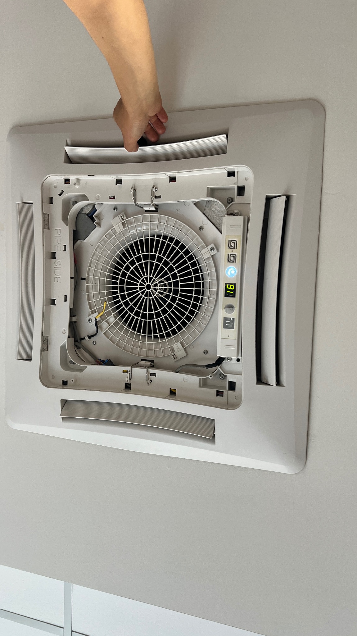 Otwarty panel sufitowej jednostki klimatyzacyjnej z widocznym wentylatorem, elektronicznym wyświetlaczem i ręką osoby regulującej ustawienia.