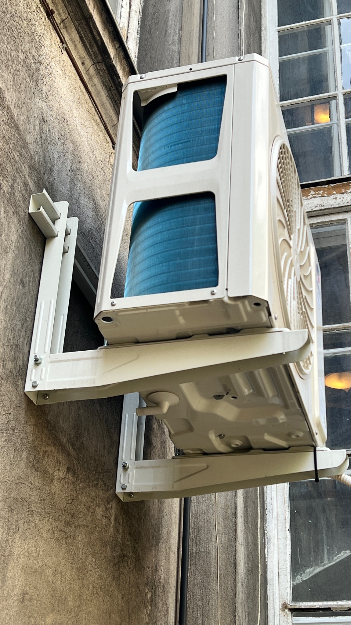 Zewnętrzna jednostka klimatyzatora zamontowana na ścianie budynku, widoczne elementy montażowe i fragment okna w tle.