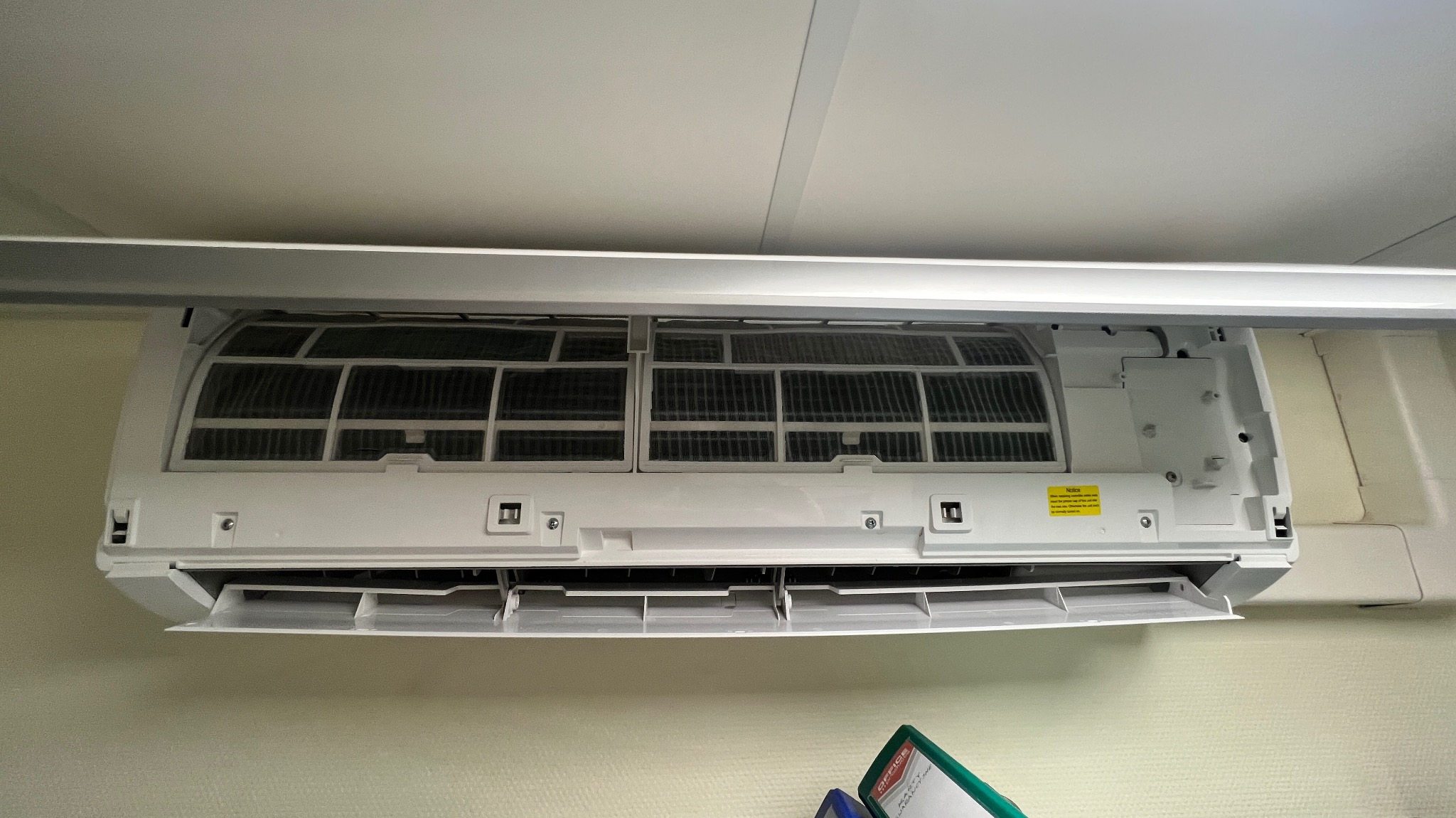 Otwarty, biały klimatyzator ścienny z widocznymi, zabrudzonymi filtrami, zamontowany pod sufitem. Żółta naklejka ostrzegawcza widoczna na obudowie.