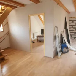 Wnętrze w trakcie prac wykończeniowych: widoczne płyty gipsowo-kartonowe na ścianach, drewniana podłoga, schody i odsłonięte belki stropowe, drabina oparta o ścianę.