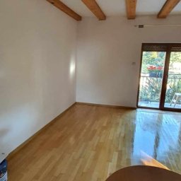 Kris - Pomalowane na jasny kolor ściany pokoju z drewnianymi belkami stropowymi, parkietem i wyjściem na balkon.