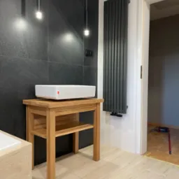 Nowoczesna łazienka z umywalką nablatową na drewnianym stoliku, czarne płytki na ścianie, grzejnik panelowy i podłoga wyłożona jasnymi panelami drewnopodobnymi.