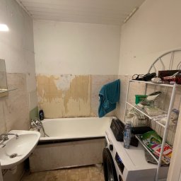 Kris - Łazienka przed remontem: widoczne stare płytki, ślady po usuniętych elementach, stara umywalka i wanna. Na wannie pralka i półka z detergentami. Widok na remontowaną łazienkę.