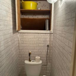 Kris - Wnętrze małej toalety z tapetą imitującą cegłę, otwartą szafką z żółtą miską i półkami z wzorzystą tapetą w tle. Nad szafką okrągła lampa.