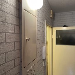 Kris - Szara ściana z cegły z metalową skrzynką z oświetleniem i przyciskami. W tle widoczne drzwi i telefon na ścianie. Ujęcie z bliska, z perspektywy lekko od dołu.