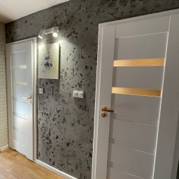 Kris - Dwa białe, nowoczesne drzwi z przeszkleniami na tle ściany z dekoracyjnym tynkiem w odcieniach szarości. Na ścianie obraz i włączniki światła. Podłoga drewniana.